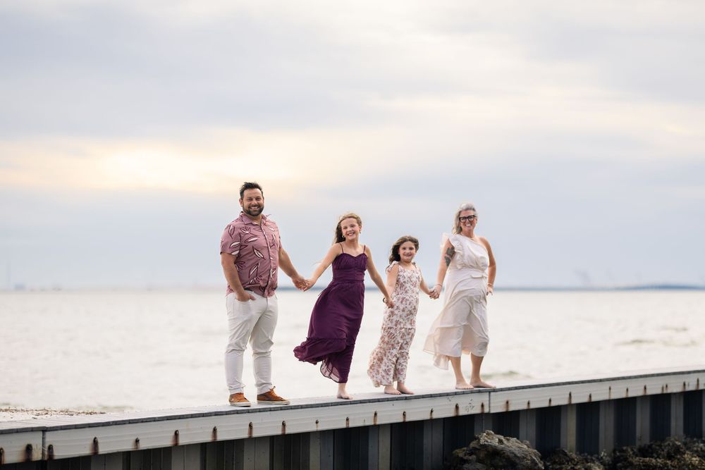A group of people in formal attire walk and laugh together on a wooden dock by the water at sunset.