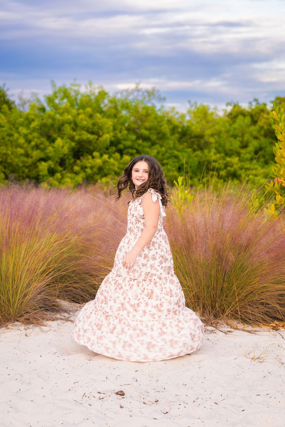 A person in a floral white gown poses on sandy ground surrounded by tall grass and green trees under blue sky.