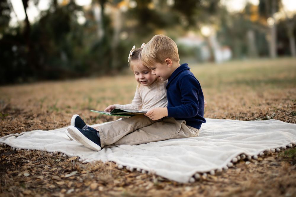 Two small children sitting on a blanket reading a book together on fallen autumn leaves.