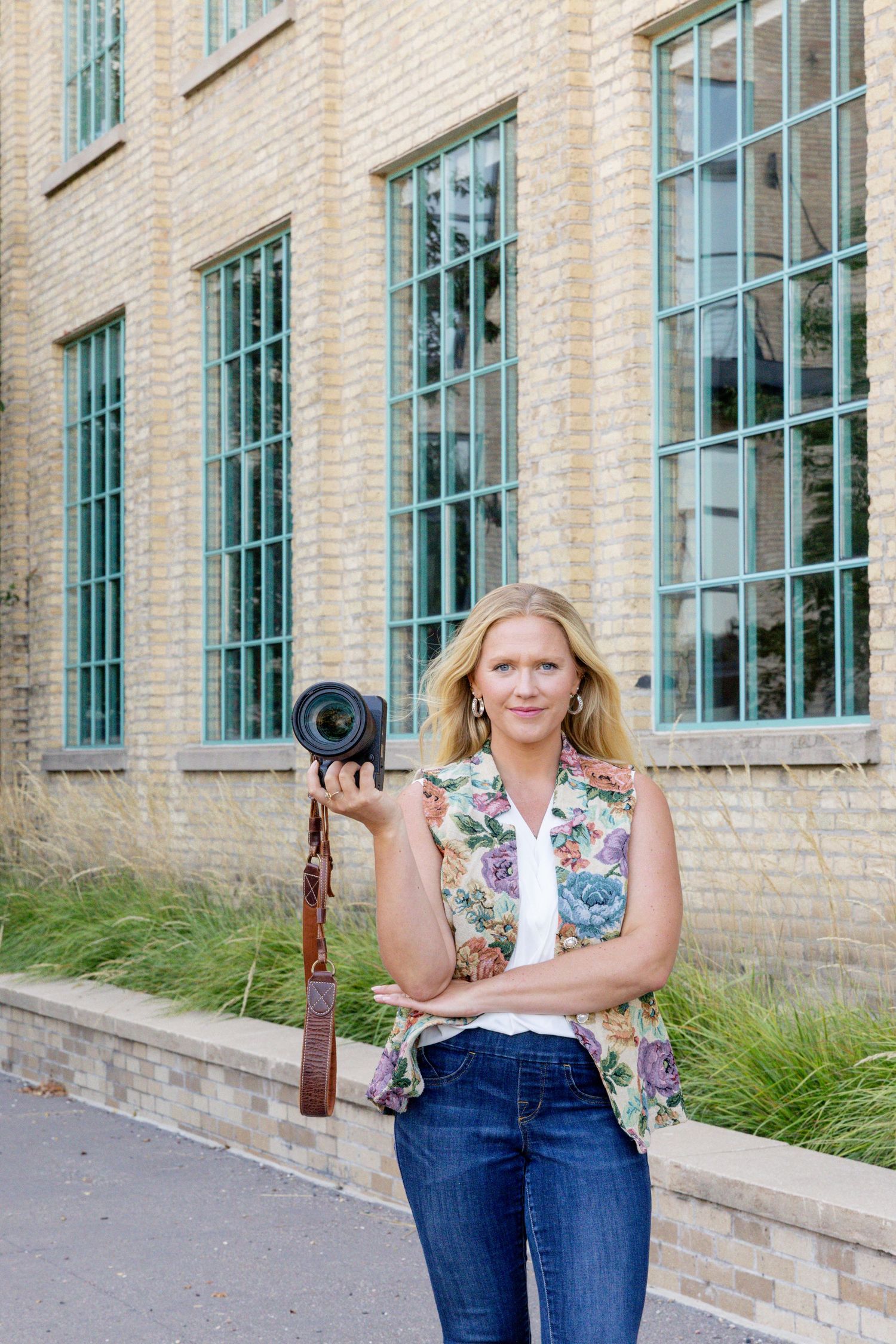 A person in a floral top and jeans poses casually against an industrial brick building with large windows.