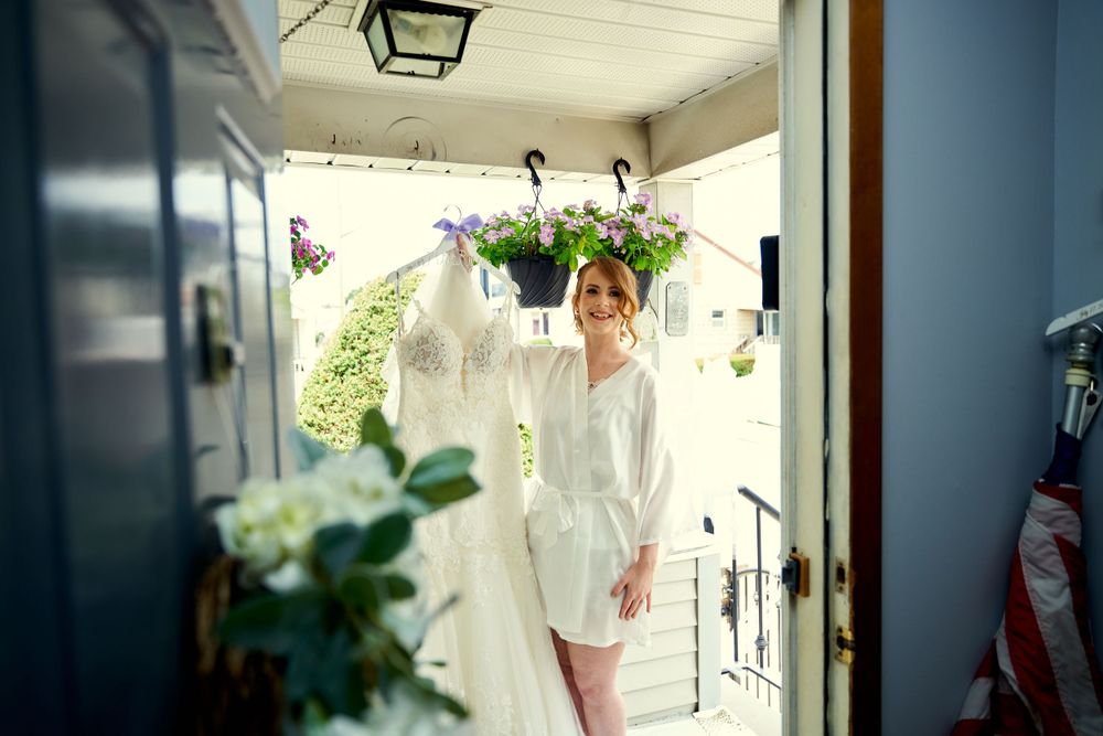 Figure in white robe stands on a sunny porch with hanging plants and white wedding dress visible.