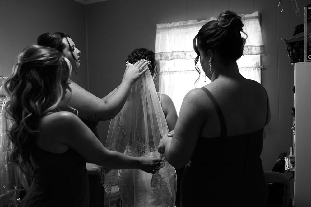 Bridesmaids help adjust a wedding veil in a backlit room during wedding preparations.