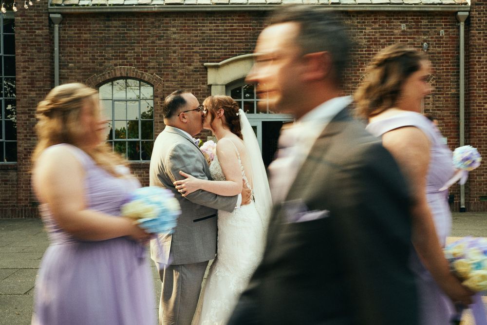 Wedding party and guests in motion during an outdoor celebration against a brick building backdrop.