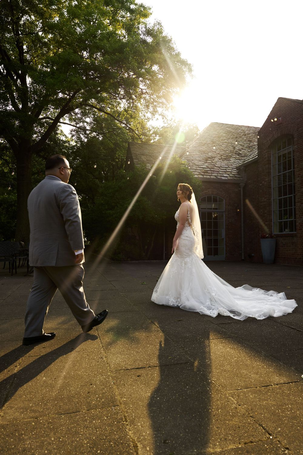 Couple walks across sunlit lawn with dramatic shadows and wedding dress train flowing behind.