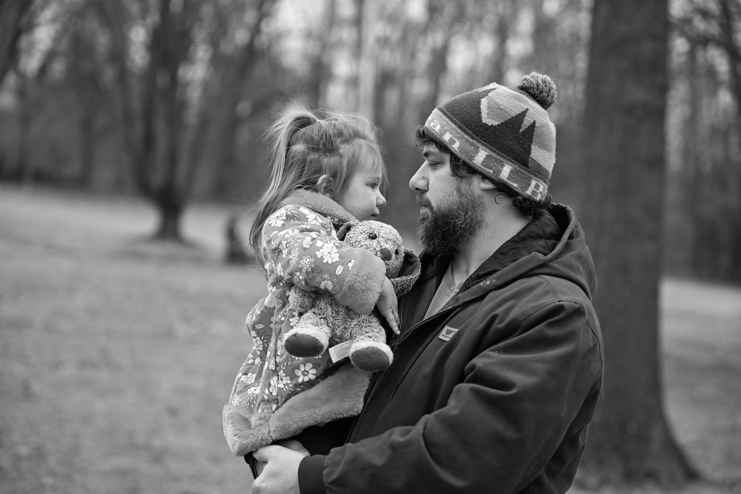 A black and white photo of Evan holding his daughter while standing in front of some trees at the park
