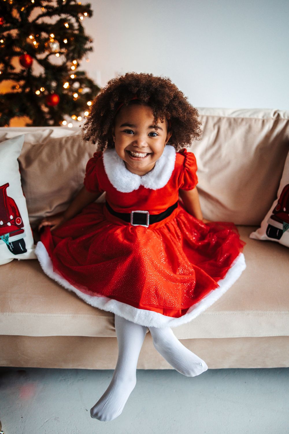 A Christmas-themed photo of a child in a red Santa dress sitting on a beige couch with a decorated tree.