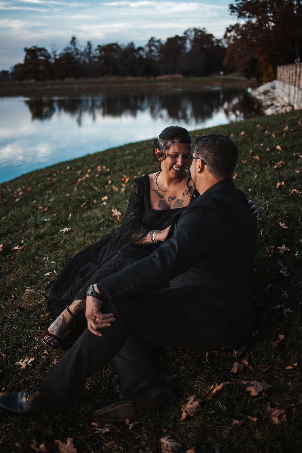 Couple relaxing by a serene lake during sunset among fallen autumn leaves.