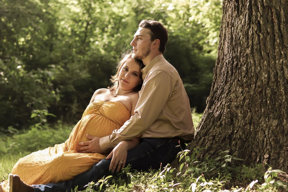 A romantic outdoor portrait of a couple resting against a tree in warm sunlight.