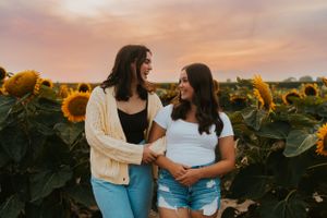 Two best friends in a sunflower field at sunset. Their arms are linked and they are smiling at each other.