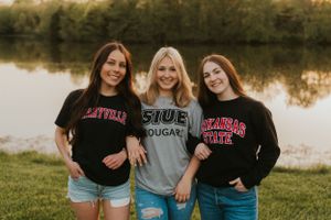 Three high school seniors wearing their chosen college t-shirts. They are smiling and holding hands while looking at the camera.