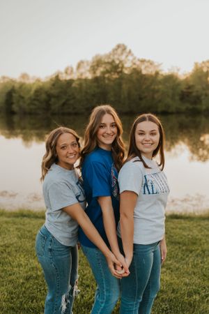 Three high school seniors wearing their chosen college t-shirts. They are smiling and holding hands while looking at the camera.