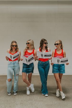 A group of girls wearing black sunglasses and red tank tops, holding newspapers and smiling at each other.