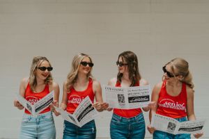 A group of girls wearing black sunglasses and red tank tops, holding newspapers and smiling at each other.