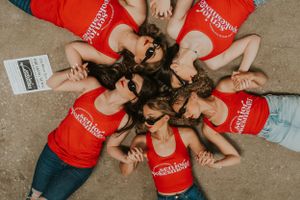 A group of girls wearing black sunglasses and red tank tops. They are laying on the ground in a circle, with their heads together.
