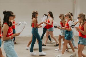 A group of young girls wearing black sunglasses and matching red tank tops. They are walking past the camera holding newspapers.