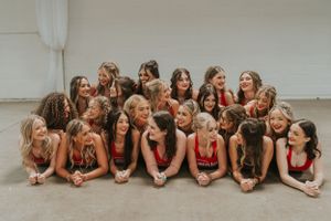A group of young ladies are smiling at each other posing for the camera. They are all wearing red tank tops.