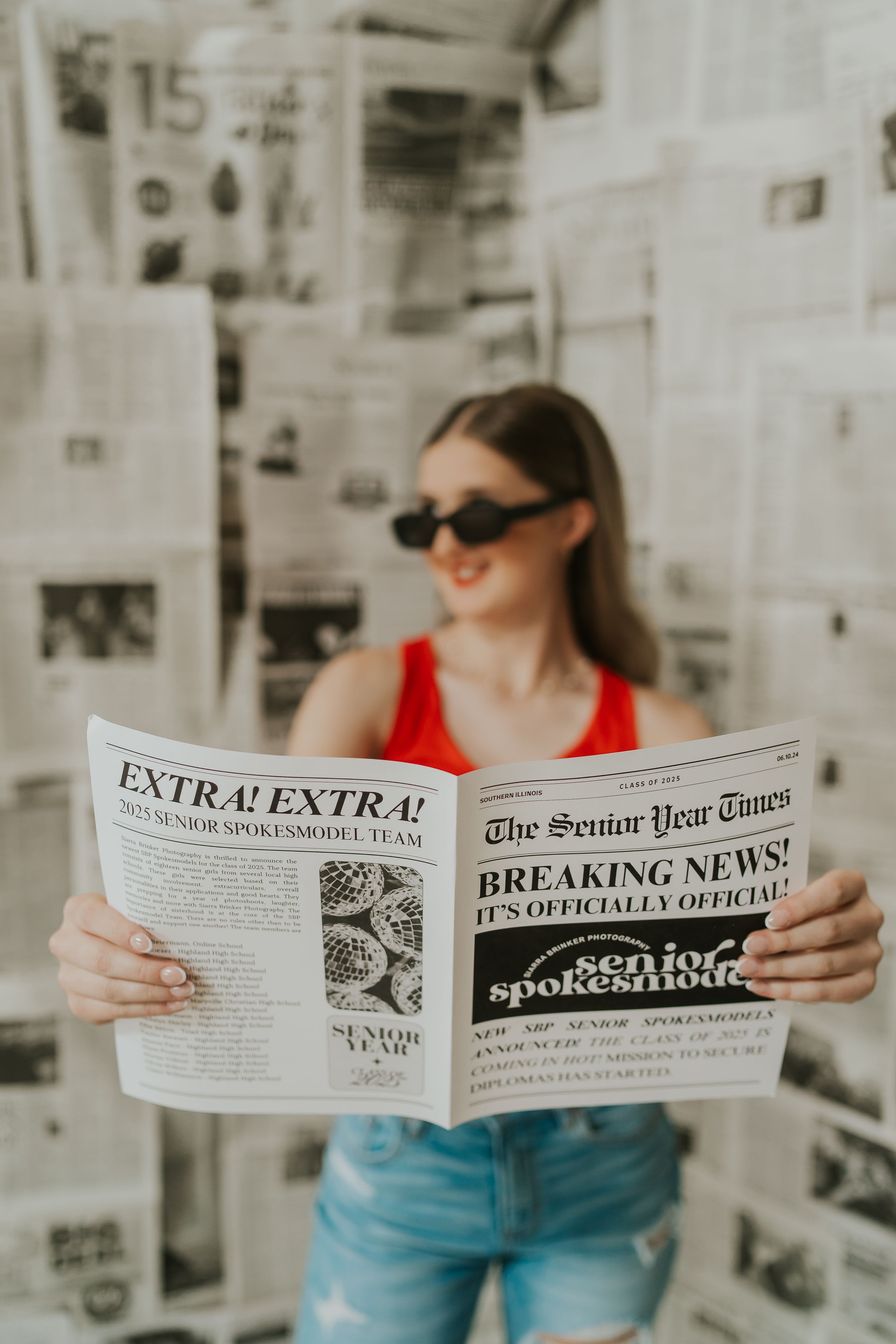 A young lady smiling off camera and holding up a newspaper.
