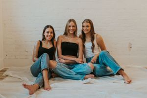Three friends sitting on the ground in a neutral room. They have glitter makeup on their faces and arms and are smiling.