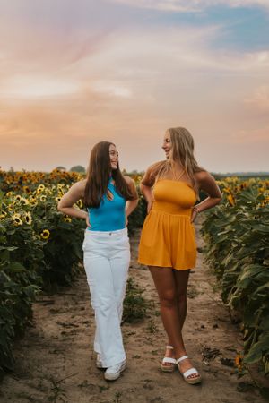 Two best friends in a sunflower field at sunset. They have their hands on their hips and are smiling at each other.