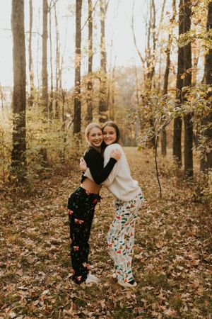 Two high school senior friends giving each other a hug. They are wearing matching christmas pajamas and are standing in the forest