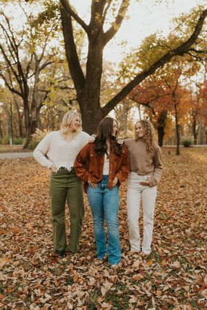 Three best friends are standing in the park amongst the autumn leaves. They have their hands in their pockets but are smiling playfully.