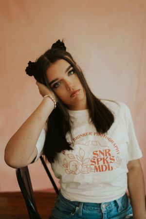 A high school senior portrait of a young lady sitting in front of a pink backdrop. She is leaning on a chair with one hand.