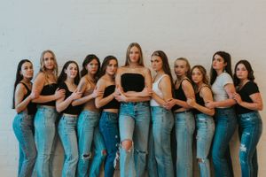 A group of high school senior girls are gathered together in a line, hugging each other. They are wearing neutral clothing and have glitter