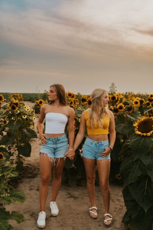 Two best friends in a sunflower field at sunset. They are holding hands and looking away from each other.