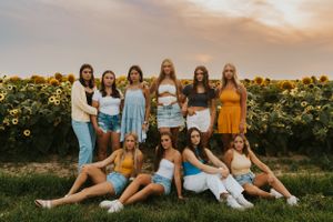 A group of high school senior girls posing for a photo at a sunflower field at sunset. They are wearing denim, yellow and white clothing.