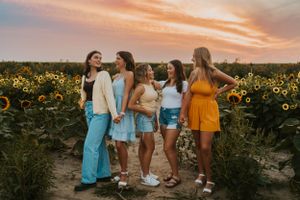 A group of high school friends standing in a sunflower patch at sunset. They are posing with one another and smiling at the camera.