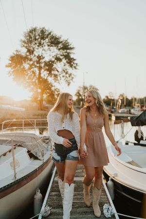 Two best friends at sunset on a boat dock, surrounded by the water and boats. They are walking toward the camera and smiling at each other.