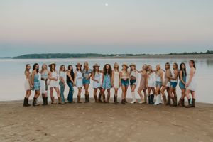 A group of young ladies wearing coastal cowgirl outfits. They are at the beach at a lake and looking happy.
