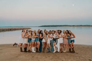 A group of young ladies huddled close and hugging. They are standing in a line on the beach at at lake during sunset and look happy.