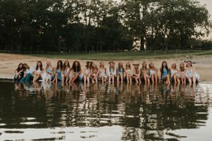 A group of young ladies on a beach at a lake. They are sitting on the edge of the water and posing for the camera.