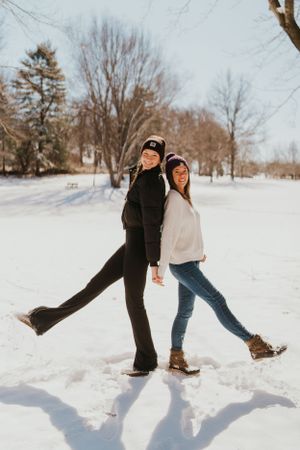 Two young women in the snow. Standing back to back, holding hands and smiling.