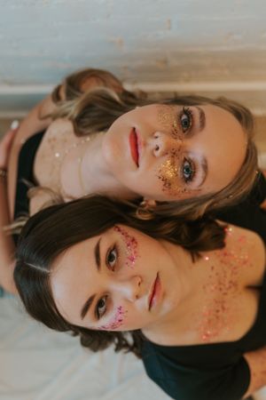 Two best friends with glitter on their face are posing for the camera. They are leaning on each other's backs.