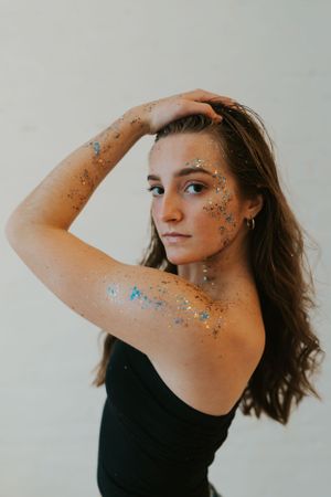 A young lady with body glitter on her arms and face, wearing a blank tank top and posing with her hands in her hair.