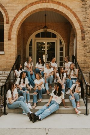 A group of high school senior girls wearing matching t-shirts sitting on the front porch of a neutral-colored brick building.