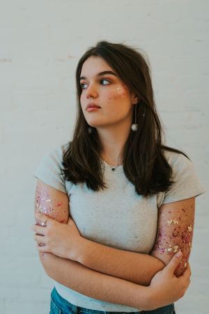A high school senior portrait of a brunette young lady with glitter makeup on her arms and face. She has her ams crossed and is looking away