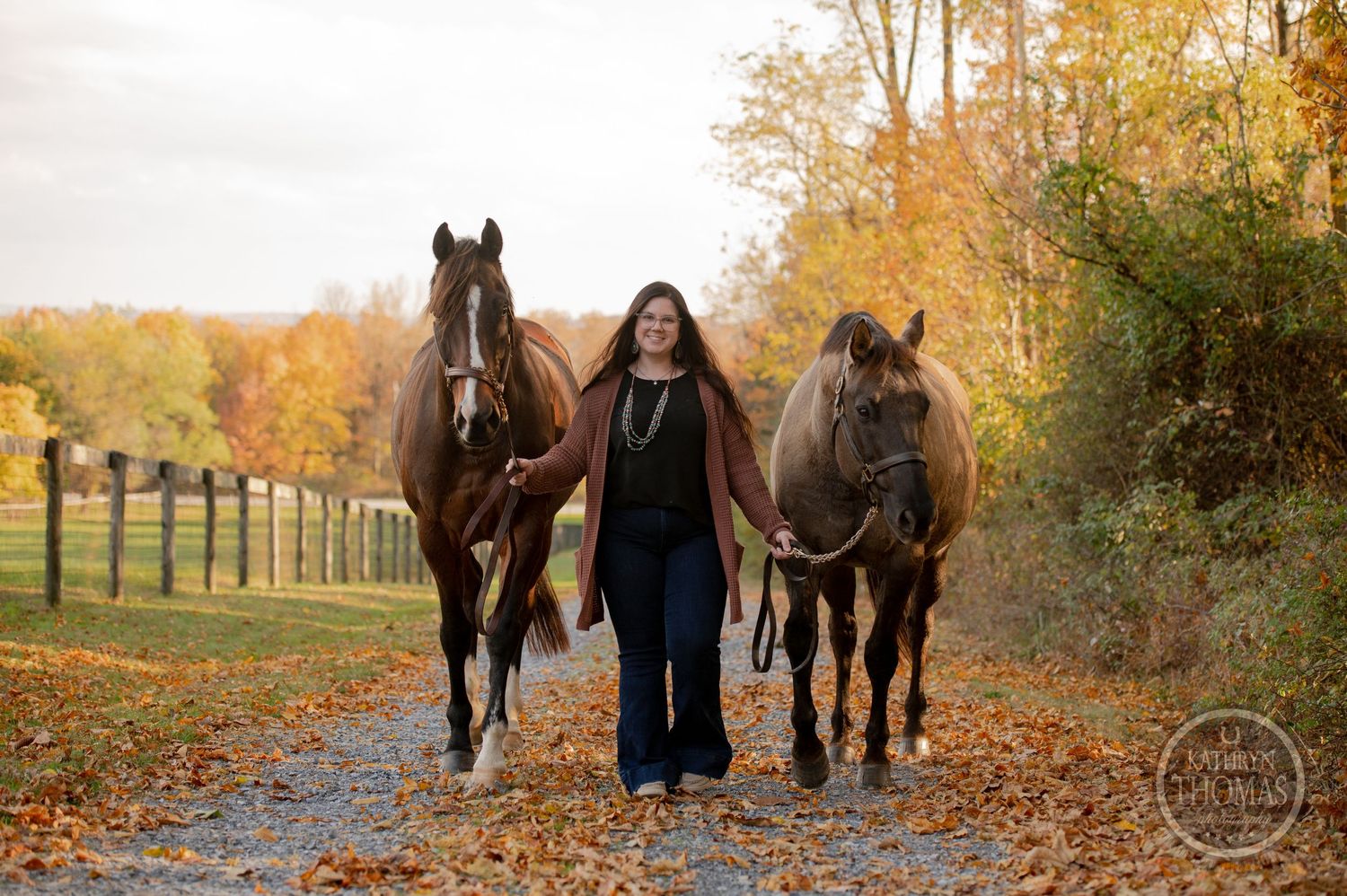 Three horses walk along a leaf-covered trail during autumn with their handler between them.
