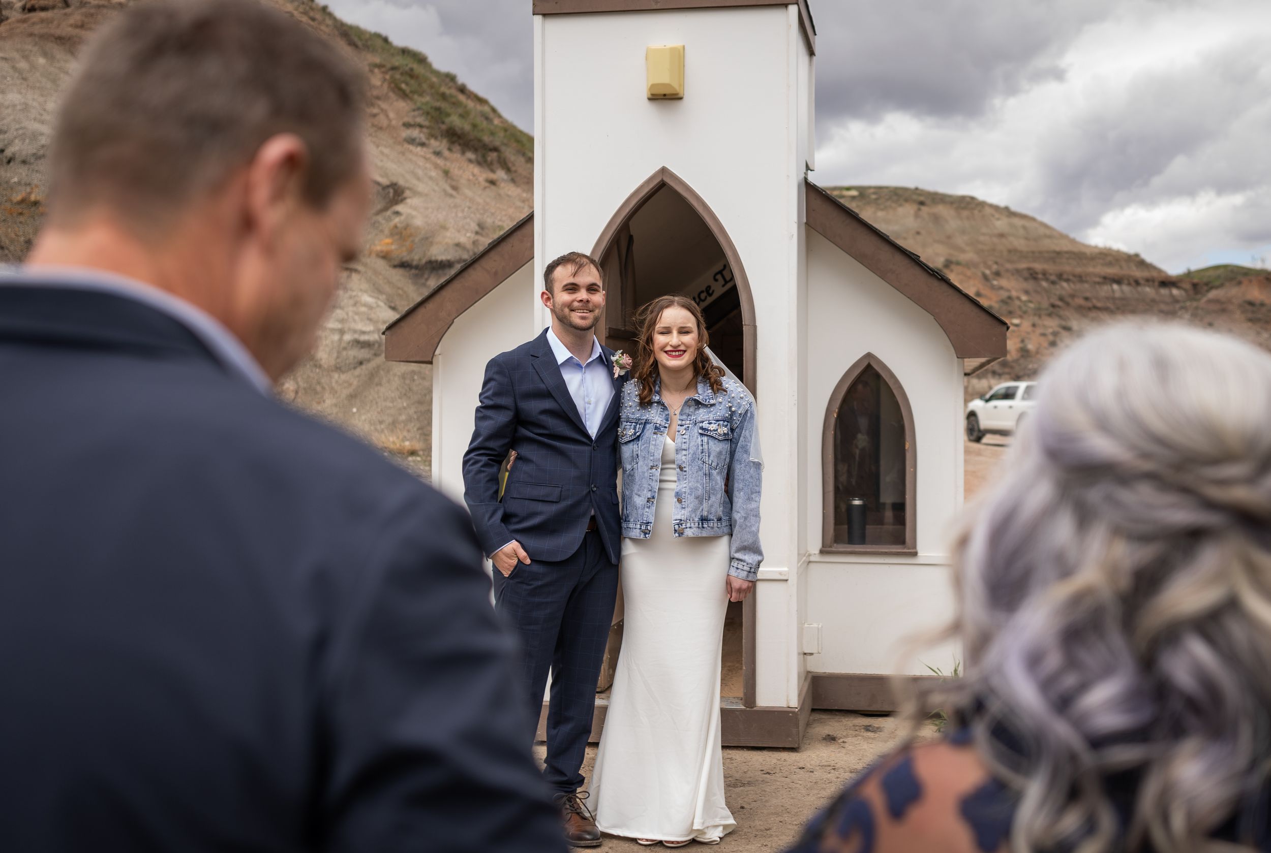 Couple poses in front of small white chapel with mountain backdrop.