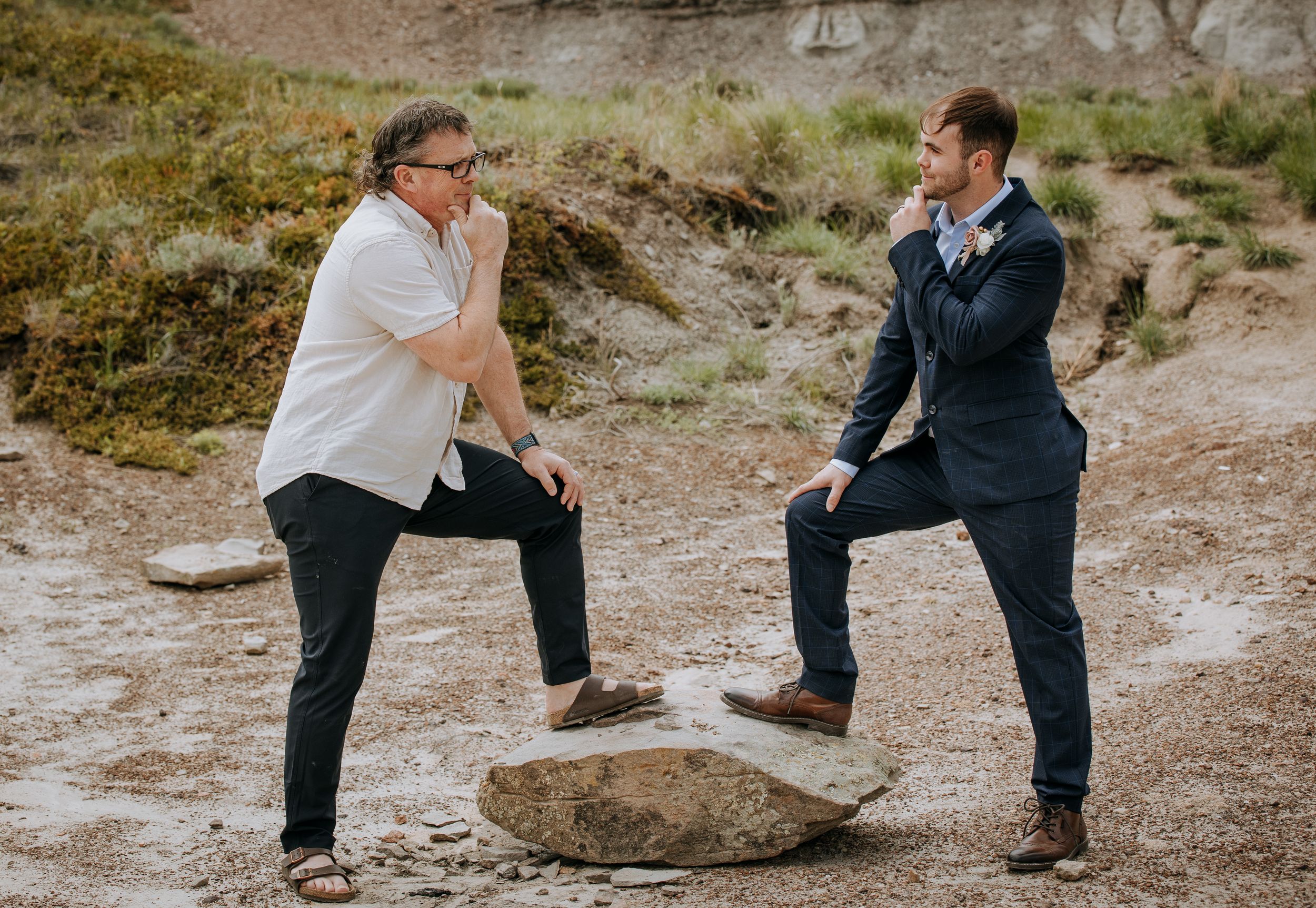 Two people strike playful poses on rock in desert landscape.