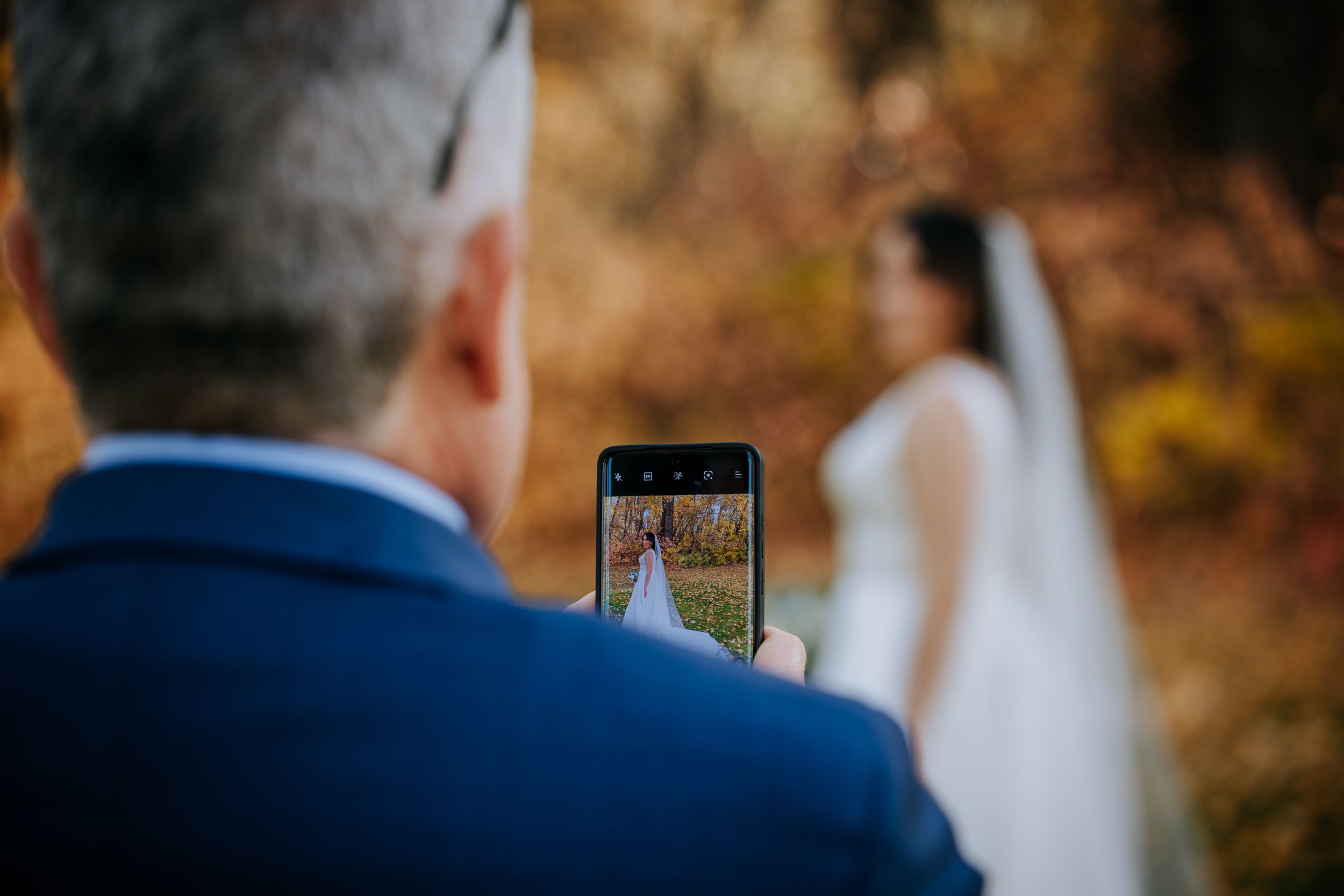 Guest captures wedding ceremony through phone screen during autumn outdoor celebration.