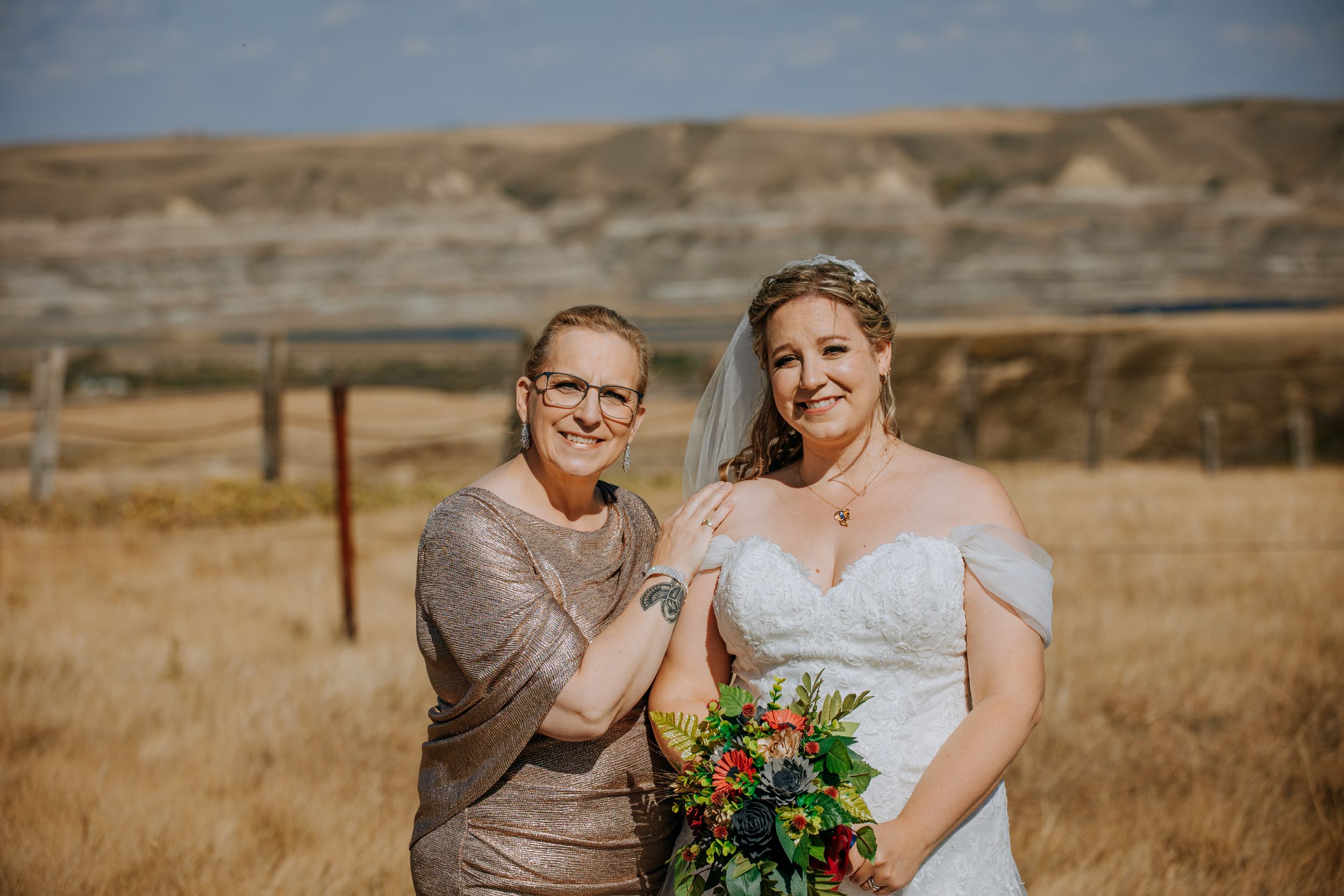 Couple poses in a rustic outdoor setting with golden fields and rugged hills in the background.