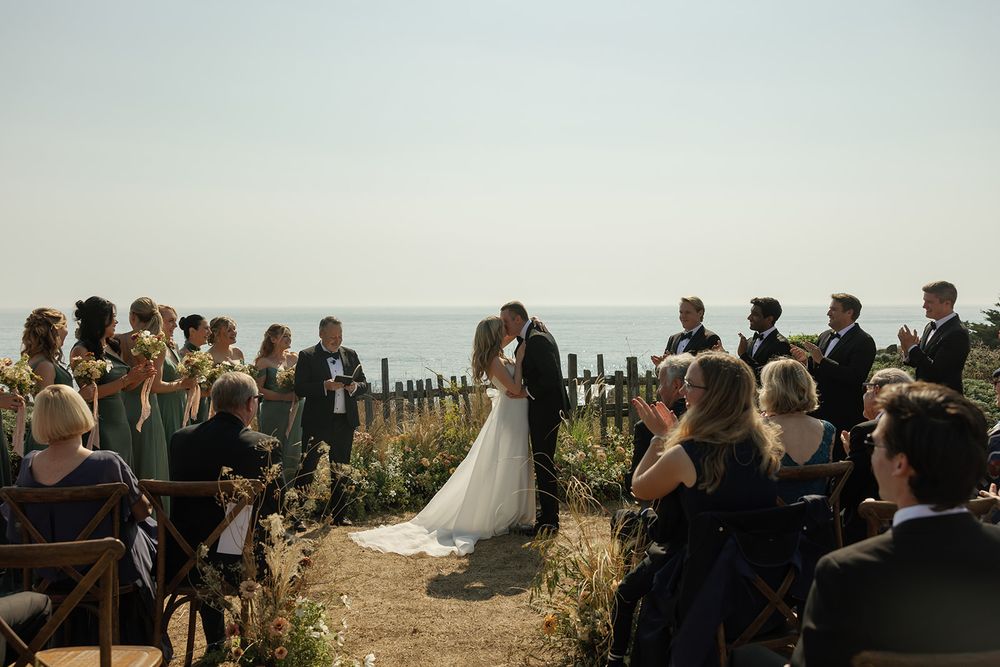 An outdoor wedding ceremony overlooking the ocean with guests seated on wooden chairs.