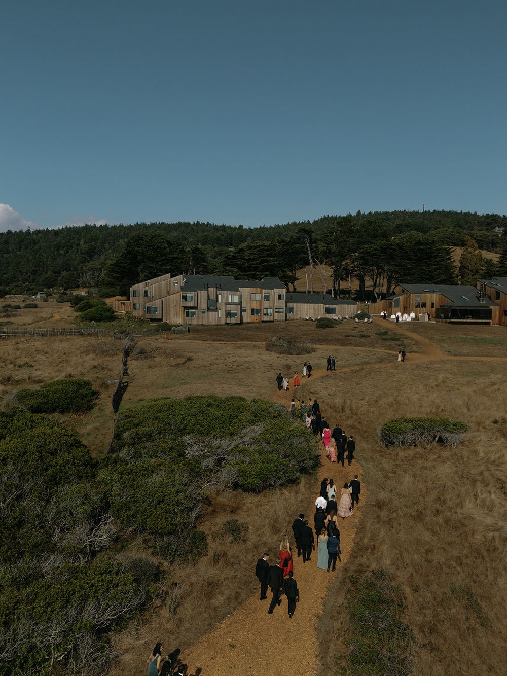 Wedding guests walking along a dirt path towards buildings on a coastal hillside.