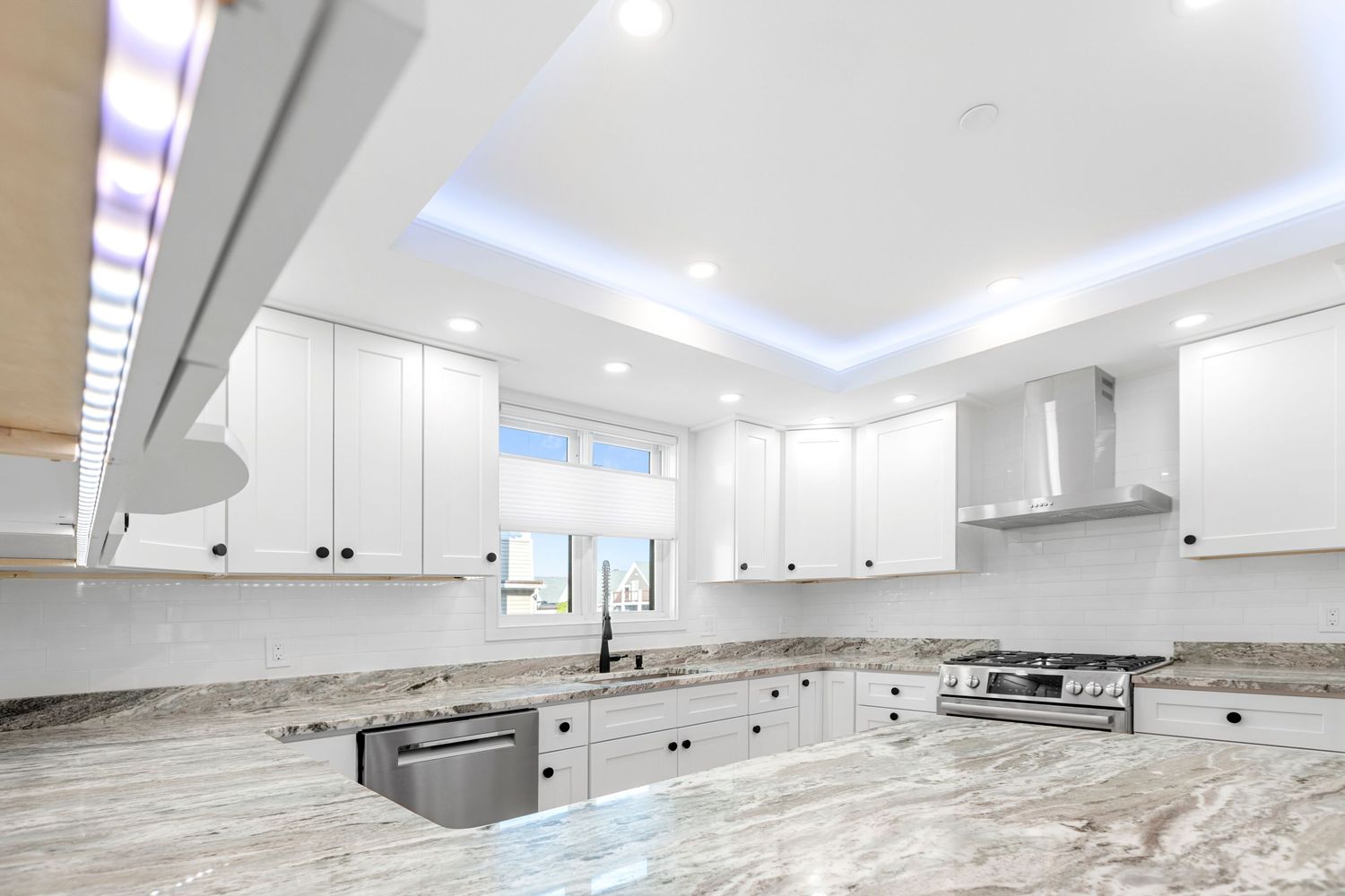 Bright white modern kitchen with marble countertops, tray ceiling with LED lighting, stainless appliances, and white cabinets.