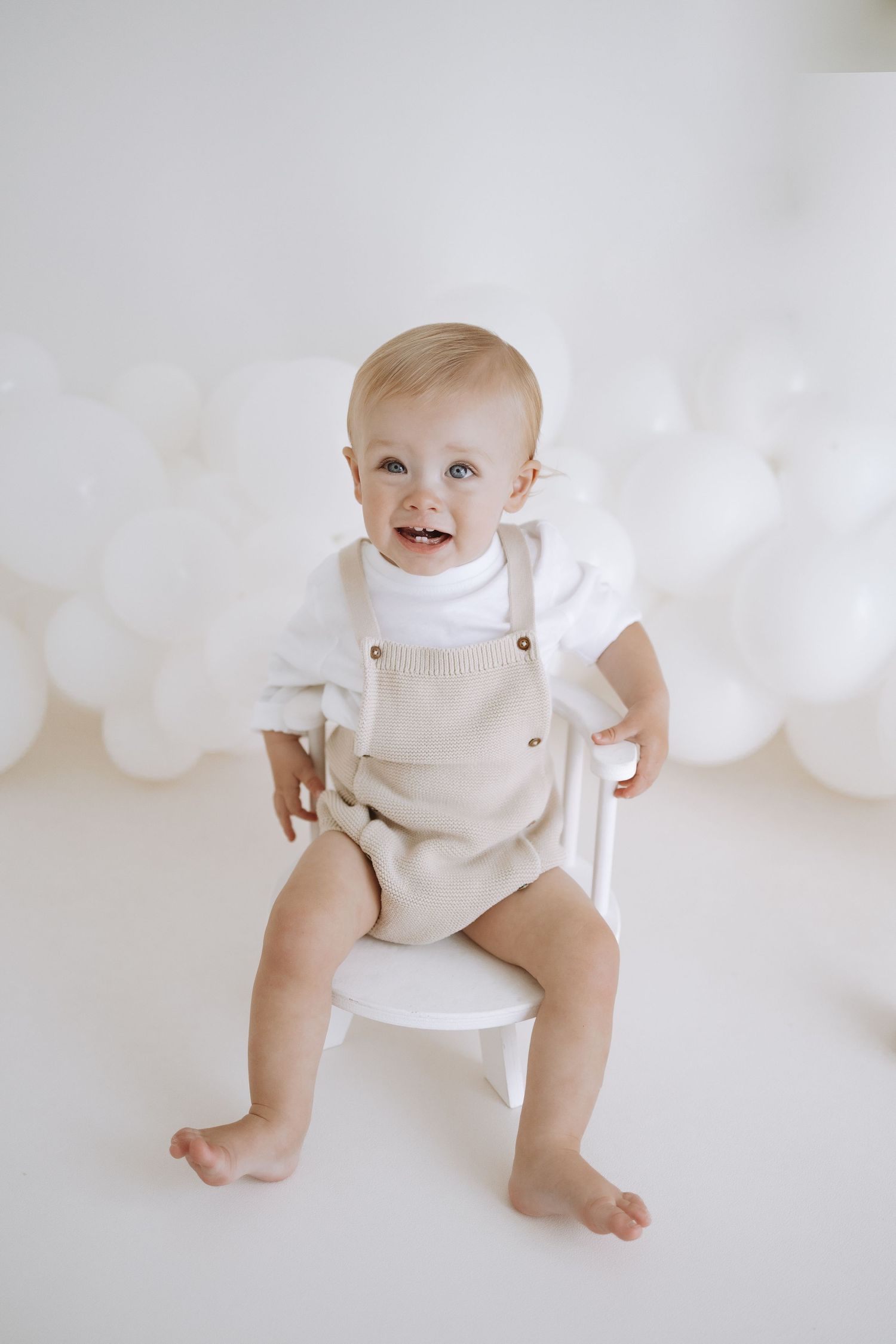 Baby in cream colored overalls and white shirt sits on white chair against light background with balloon decorations.