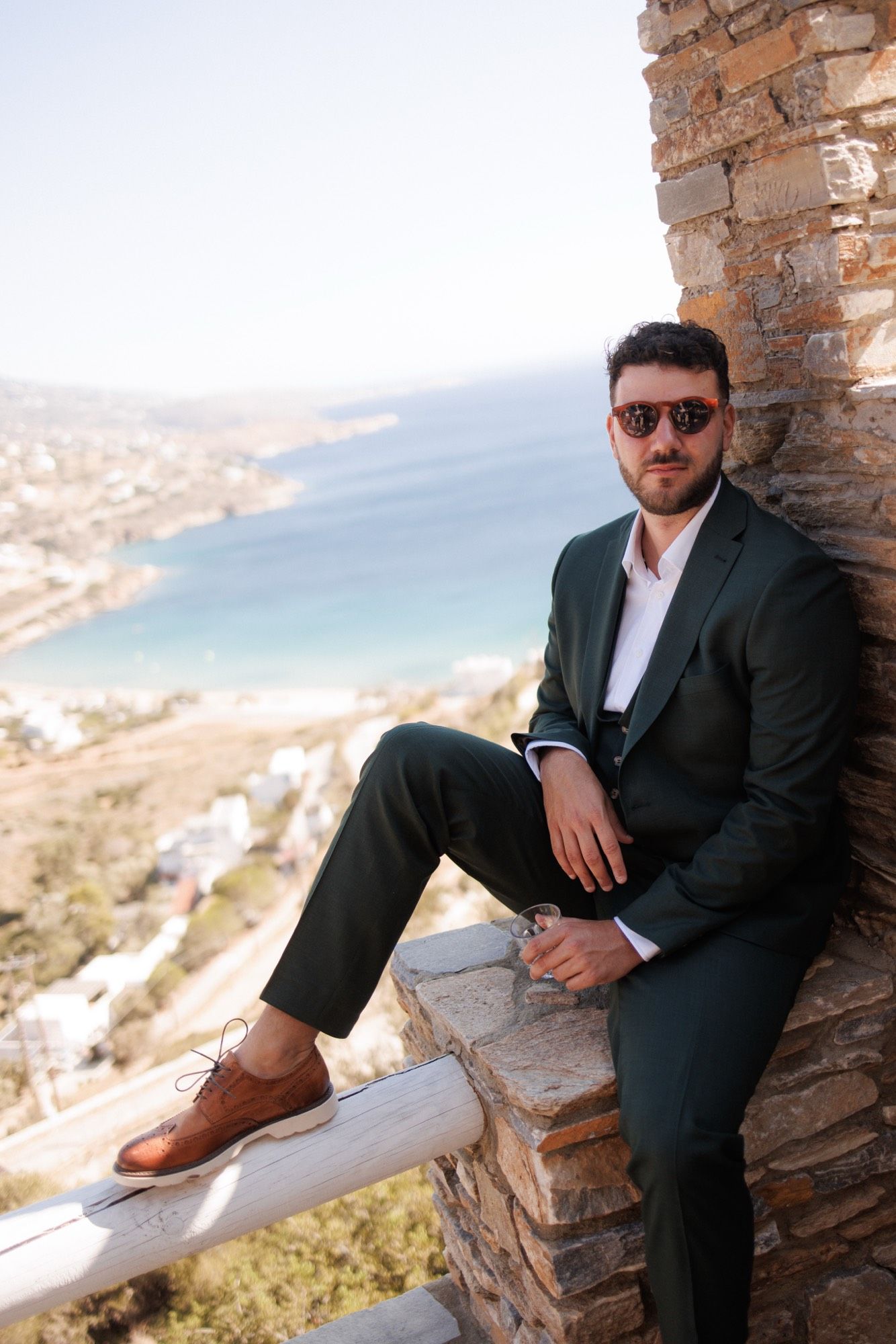 Man in black suit and sunglasses poses casually on stone wall overlooking coastal Mediterranean landscape.