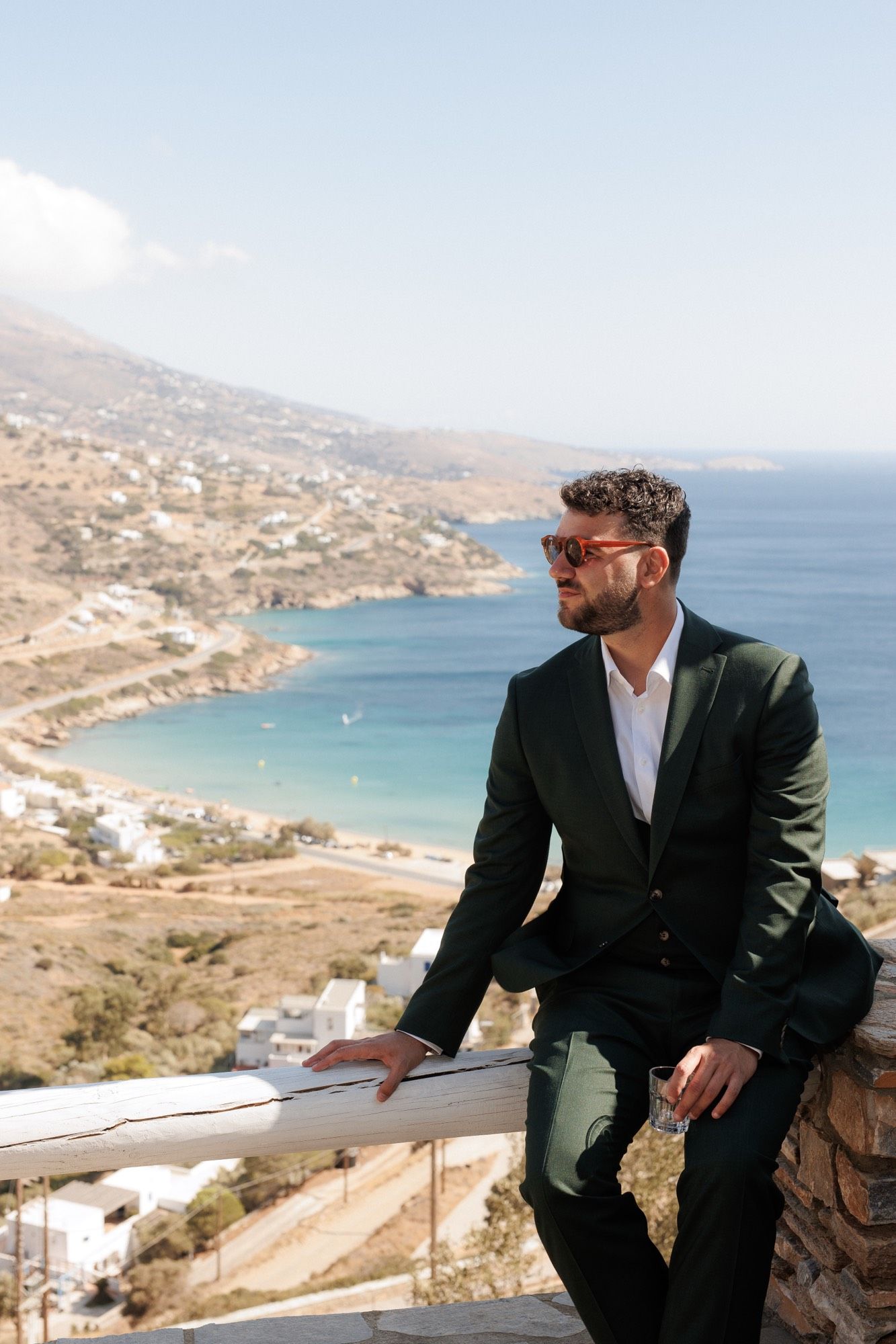 A person in a dark suit jacket gazes out at a scenic coastal Mediterranean landscape with turquoise waters below.